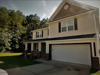 A suburban two-story house with a gabled roof and black shutters. The exterior has light-colored siding and brick accents, with a spacious front porch supported by white columns. A two-car garage is present, and the house is surrounded by lush green grass and trees.