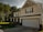 A suburban two-story house with a gabled roof and black shutters. The exterior has light-colored siding and brick accents, with a spacious front porch supported by white columns. A two-car garage is present, and the house is surrounded by lush green grass and trees.