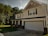 A suburban two-story house with a gabled roof and black shutters. The exterior has light-colored siding and brick accents, with a spacious front porch supported by white columns. A two-car garage is present, and the house is surrounded by lush green grass and trees.