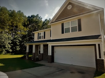 A suburban two-story house with a gabled roof and black shutters. The exterior has light-colored siding and brick accents, with a spacious front porch supported by white columns. A two-car garage is present, and the house is surrounded by lush green grass and trees.