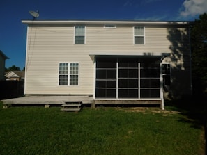 A two-story house with a beige exterior and multiple windows is visible. The house features a screened porch attached to a wooden deck, overlooking a green lawn. A satellite dish is mounted on the roof.
