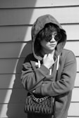 A person in a hoodie stands against a wooden wall, wearing large round sunglasses and holding a stylish purse with a chain strap. The composition is in black and white, emphasizing the textures of the clothing and wall.