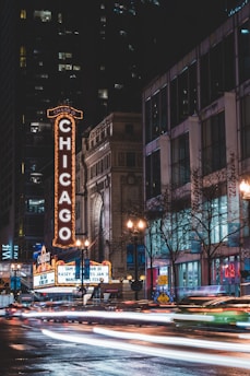 A vibrant city street scene at night featuring the iconic Chicago Theater sign illuminated in bright lights. Surrounding buildings rise tall with numerous windows, and the streets are bustling with blurred car lights, indicating movement. The atmosphere is lively with lit-up billboards and signage, and leafless trees line the street.