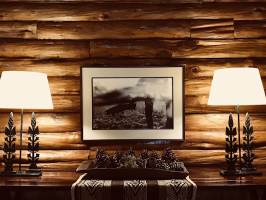 A cozy log cabin interior features a wooden wall with a framed black-and-white photograph at the center. The photograph is flanked by two identical table lamps with decorative stands resembling tree branches. Below the photograph, a wooden table holds a decorative arrangement of pine cones on a woven runner.