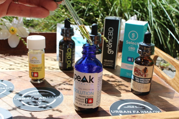 Various bottles and packaging of CBD products are arranged on a wooden surface outdoors. A hand holds a dropper with liquid above a blue glass bottle labeled 'PEAK Extracts Full Spectrum Solution'. The surrounding products include softgels, tinctures, and CBD oil from different brands. Urban Farmacy stickers are visible on the table, and there is foliage in the background.