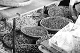 A variety of baskets filled with assorted dried goods and spices are displayed. The baskets contain items such as dried vegetables, nuts, and seeds in a market setting. A person’s arm is visible, possibly indicating a market vendor or a customer reaching into the baskets.