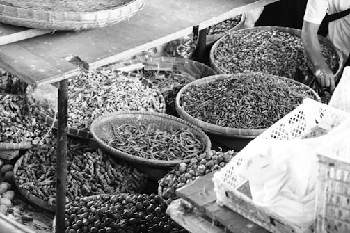 A variety of baskets filled with assorted dried goods and spices are displayed. The baskets contain items such as dried vegetables, nuts, and seeds in a market setting. A person’s arm is visible, possibly indicating a market vendor or a customer reaching into the baskets.