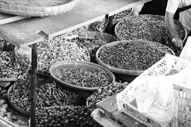 A variety of baskets filled with assorted dried goods and spices are displayed. The baskets contain items such as dried vegetables, nuts, and seeds in a market setting. A person’s arm is visible, possibly indicating a market vendor or a customer reaching into the baskets.