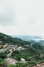 A lush, green mountainous landscape overlooks a coastal town dotted with small houses and winding roads. The scene is enveloped in a tranquil atmosphere with dense vegetation covering the hills. In the distance, water bodies are visible under a cloudy sky, creating a serene backdrop.
