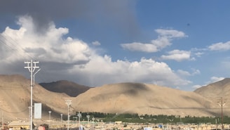 A landscape featuring arid mountains under a partly cloudy sky with a mix of sunlight and shadow. Power lines and poles are visible in the foreground, indicating a connection to human habitation.