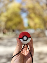 A hand is holding a round device resembling a Poké Ball, featuring a red upper half and a white lower half with a black horizontal band and a button in the center. The background is blurred, showing shades of yellow and green, suggesting an outdoor setting with trees.