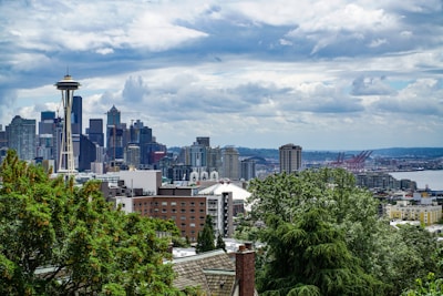 A city skyline with a prominent observation tower, likely the Space Needle, surrounded by various skyscrapers and buildings. In the foreground, there are lush green trees, and in the background, there is a waterfront with industrial structures, possibly cranes, under a partly cloudy sky.