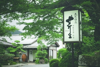 A tranquil setting with a traditional Japanese building surrounded by lush green foliage. A wooden sign with Japanese characters is prominently displayed in the foreground. The landscape is manicured, with carefully trimmed hedges and potted plants placed strategically along a gravel path.
