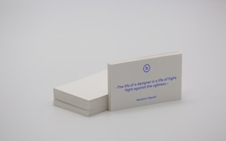 A stack of white business cards is placed on a plain surface, with one card prominently displayed at the front. The card features a quote in blue text that reads, 'The life of a designer is a life of fight: fight against the ugliness.' The name Massimo Vignelli is printed below the quote. The overall scene is minimalist and simple.