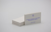 A stack of white business cards is placed on a plain surface, with one card prominently displayed at the front. The card features a quote in blue text that reads, 'The life of a designer is a life of fight: fight against the ugliness.' The name Massimo Vignelli is printed below the quote. The overall scene is minimalist and simple.