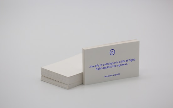A stack of white business cards is placed on a plain surface, with one card prominently displayed at the front. The card features a quote in blue text that reads, 'The life of a designer is a life of fight: fight against the ugliness.' The name Massimo Vignelli is printed below the quote. The overall scene is minimalist and simple.