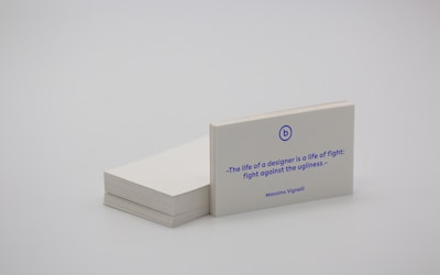 A stack of white business cards is placed on a plain surface, with one card prominently displayed at the front. The card features a quote in blue text that reads, 'The life of a designer is a life of fight: fight against the ugliness.' The name Massimo Vignelli is printed below the quote. The overall scene is minimalist and simple.
