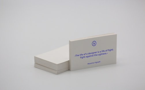 A stack of white business cards is placed on a plain surface, with one card prominently displayed at the front. The card features a quote in blue text that reads, 'The life of a designer is a life of fight: fight against the ugliness.' The name Massimo Vignelli is printed below the quote. The overall scene is minimalist and simple.