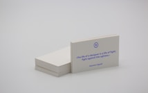 A stack of white business cards is placed on a plain surface, with one card prominently displayed at the front. The card features a quote in blue text that reads, 'The life of a designer is a life of fight: fight against the ugliness.' The name Massimo Vignelli is printed below the quote. The overall scene is minimalist and simple.