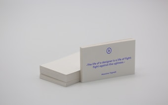 A stack of white business cards is placed on a plain surface, with one card prominently displayed at the front. The card features a quote in blue text that reads, 'The life of a designer is a life of fight: fight against the ugliness.' The name Massimo Vignelli is printed below the quote. The overall scene is minimalist and simple.