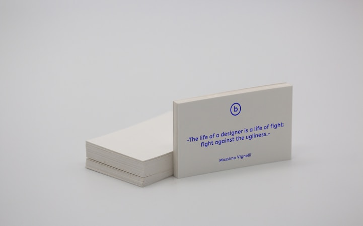 A stack of white business cards is placed on a plain surface, with one card prominently displayed at the front. The card features a quote in blue text that reads, 'The life of a designer is a life of fight: fight against the ugliness.' The name Massimo Vignelli is printed below the quote. The overall scene is minimalist and simple.