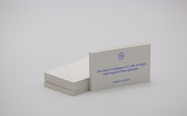 A stack of white business cards is placed on a plain surface, with one card prominently displayed at the front. The card features a quote in blue text that reads, 'The life of a designer is a life of fight: fight against the ugliness.' The name Massimo Vignelli is printed below the quote. The overall scene is minimalist and simple.