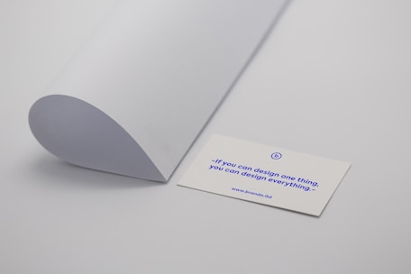 A minimalistic scene with a curved sheet of white paper positioned next to a business card. The business card displays a motivational quote in blue text: 'If you can design one thing, you can design everything.' The card rests on a plain light grey surface.
