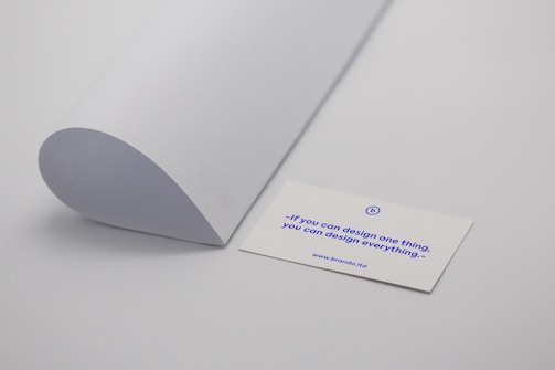 A minimalistic scene with a curved sheet of white paper positioned next to a business card. The business card displays a motivational quote in blue text: 'If you can design one thing, you can design everything.' The card rests on a plain light grey surface.