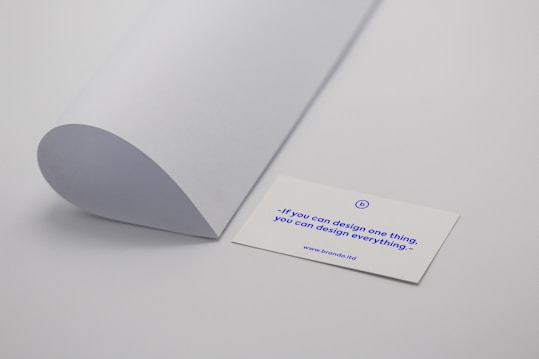 A minimalistic scene with a curved sheet of white paper positioned next to a business card. The business card displays a motivational quote in blue text: 'If you can design one thing, you can design everything.' The card rests on a plain light grey surface.