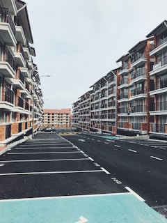 Two rows of modern, low-rise apartment buildings with multiple balconies face each other, creating a symmetrical view. The buildings have a brick facade and are separated by a wide, empty parking lot with clearly marked parking spaces. The sky above is overcast, adding a muted tone to the scene.