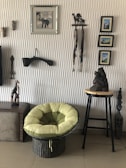 An interior scene featuring a wicker chair with plush green cushions, set against a textured wall adorned with various decorative items. The wall displays framed pictures, wooden carvings of animals, and a small stool with a carved statue. The floor appears to be tiled.