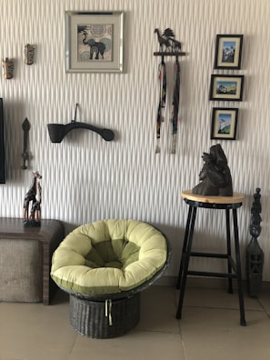 An interior scene featuring a wicker chair with plush green cushions, set against a textured wall adorned with various decorative items. The wall displays framed pictures, wooden carvings of animals, and a small stool with a carved statue. The floor appears to be tiled.
