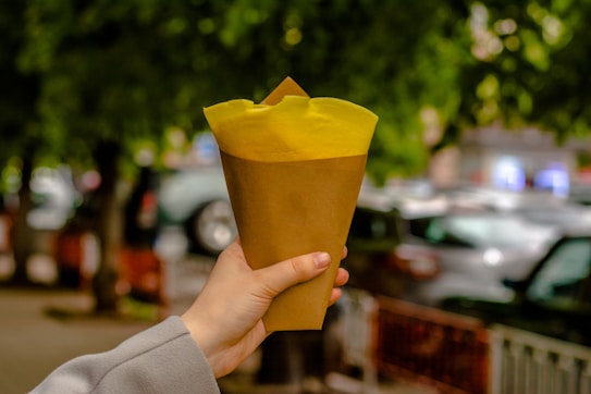 A hand is holding a brown paper cone with bright yellow tissue or cloth peeking out from the top. The background is blurred, showing parked cars and green trees, suggesting an outdoor setting.