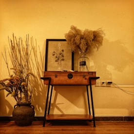 A cozy and warm interior scene featuring a wooden table against a yellowish wall. On the table, there is a framed artwork of flowers, a large glass vase filled with dried plants, and a decorative wooden box. To the left, a dark ceramic pot holds tall dried branches, adding a rustic touch. The lighting casts soft shadows, enhancing the warm ambiance.
