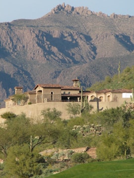 A rustic villa with stucco walls and terracotta roofs nestled in a desert landscape at the base of a rocky mountain range. Cacti and desert flora surround the property, indicating an arid environment.
