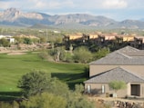 A scenic view of a golf course with well-maintained green lawns, surrounded by desert landscape. In the background, there are residential buildings styled with southwestern architecture, and beyond them are rugged mountains under a partly cloudy sky. Tall cacti and various desert shrubs dot the landscape.