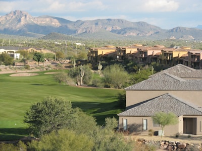 A scenic view of a golf course with well-maintained green lawns, surrounded by desert landscape. In the background, there are residential buildings styled with southwestern architecture, and beyond them are rugged mountains under a partly cloudy sky. Tall cacti and various desert shrubs dot the landscape.
