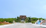 A small thatched-roof chapel surrounded by greenery stands at the end of a paved pathway. The path is flanked by colorful grave markers and crosses, with palm trees adding to the tropical feel. A person is seen walking on the pathway towards the chapel.