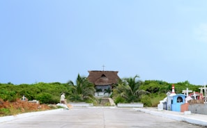A small thatched-roof chapel surrounded by greenery stands at the end of a paved pathway. The path is flanked by colorful grave markers and crosses, with palm trees adding to the tropical feel. A person is seen walking on the pathway towards the chapel.