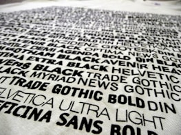 A collection of various fonts and typefaces printed in black ink on a white background, with names like Helvetica, Trade Gothic, and DIN. The text is bold and arranged in a scattered manner, showcasing different font weights and styles.