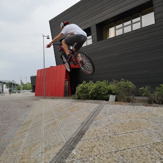 A person on a BMX bike performs an aerial trick near a modern building with dark panels and red accents. The environment features some greenery and urban landscape elements, with another bicyclist visible in the background.