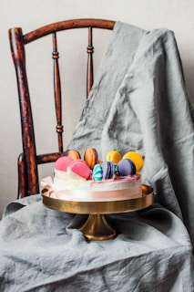 A decorative cake topped with colorful macarons is placed on a gold cake stand. This setup is positioned on a wooden chair, with a draped gray cloth serving as a backdrop, creating a rustic and elegant presentation.