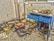 A cluttered and dusty workspace with a blue desk featuring open drawers. On and around the desk, there are various tools, electronic parts, cardboard boxes, and other assorted items scattered across the concrete floor. The walls are worn and the area looks neglected, suggesting a sense of disarray and abandonment.
