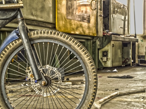 A workshop or industrial setting featuring a close-up view of a bicycle wheel in the foreground. The wheel is black with tire treads and spokes, and behind it is machinery that appears aged and metallic. Various tools and debris are scattered on the floor, and the overall environment looks worn and used.