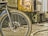 A workshop or industrial setting featuring a close-up view of a bicycle wheel in the foreground. The wheel is black with tire treads and spokes, and behind it is machinery that appears aged and metallic. Various tools and debris are scattered on the floor, and the overall environment looks worn and used.