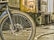 A workshop or industrial setting featuring a close-up view of a bicycle wheel in the foreground. The wheel is black with tire treads and spokes, and behind it is machinery that appears aged and metallic. Various tools and debris are scattered on the floor, and the overall environment looks worn and used.