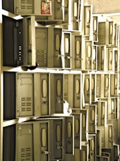 A series of metal lockers, many of which have their doors open. The lockers are arranged in a grid-like pattern against the wall, and there is a packet of snacks inside one of the open lockers. The lighting creates a warm and slightly vintage atmosphere.