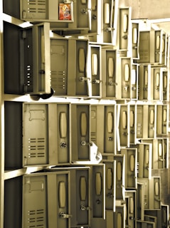 A series of metal lockers, many of which have their doors open. The lockers are arranged in a grid-like pattern against the wall, and there is a packet of snacks inside one of the open lockers. The lighting creates a warm and slightly vintage atmosphere.