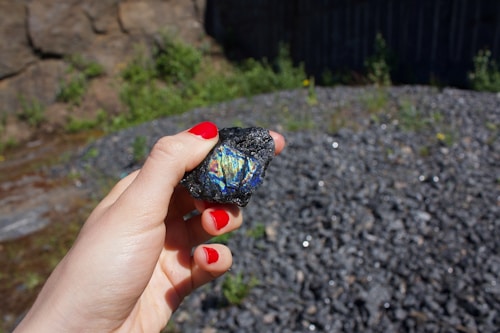 A hand with red painted nails holds a dark rock with a colorful, iridescent patch in its center. The background consists of a rocky terrain with some greenery and what appears to be a stone wall or natural rock formation.