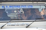 A man inside a vehicle with his hands pressed against the windshield. He appears to be making a playful or surprised expression. The vehicle has a sign with the name 'K. S. Motors Pvt. Ltd.' on the windshield and is a Mahindra model. Another person is seated next to the man inside the vehicle.